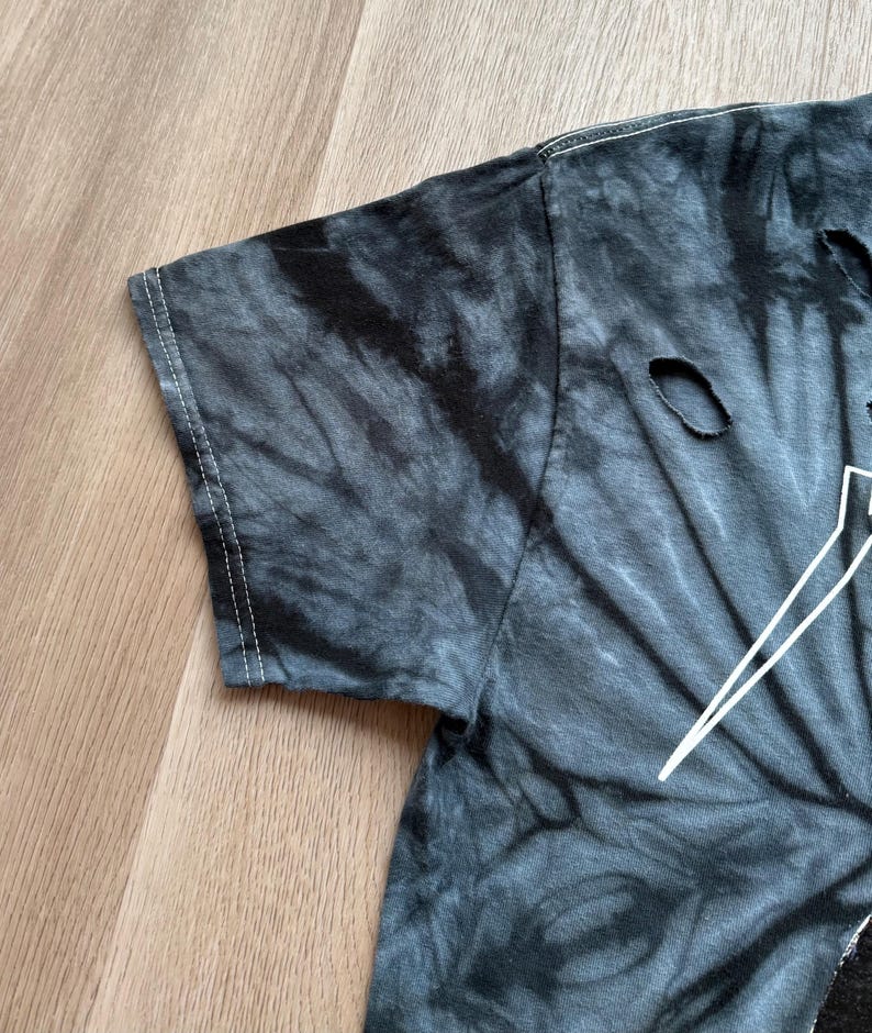 Może przedstawiać: Zbliżenie na koszulkę tie-dye w odcieniach szarości i czerni. Koszulka ma postarzany wygląd z kilkoma małymi dziurami. Białe szwy są widoczne wzdłuż szw&oacute;w rękawa.