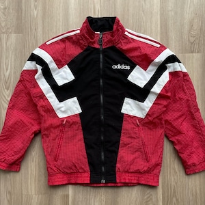 Puede incluir: Una chaqueta cortavientos roja y negra con patrones geométricos blancos. La chaqueta tiene cierre de cremallera y un logotipo que dice "adidas" en el pecho izquierdo.
