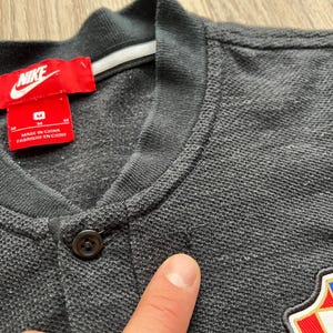 May include: A gray polo shirt with a black button and a Nike logo on the left chest. The shirt has a white tag with the text "Made in China Fabrique en Chine" and the size "M".