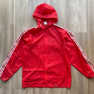 Op de afbeelding: Een rode Adidas jas met capuchon en witte strepen op de mouwen. De jas heeft een volledige ritssluiting en een klein Adidas-logo bij de zoom. De jas is gemaakt van lichtgewicht materiaal en heeft een capuchon.