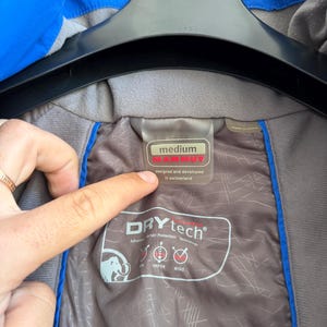 May include: A blue jacket with a grey interior lining featuring the Mammut brand logo and text. The label indicates a size medium, designed and developed in Switzerland. The interior fabric displays Drytech technology branding with icons for rain, vapor, and wind protection.