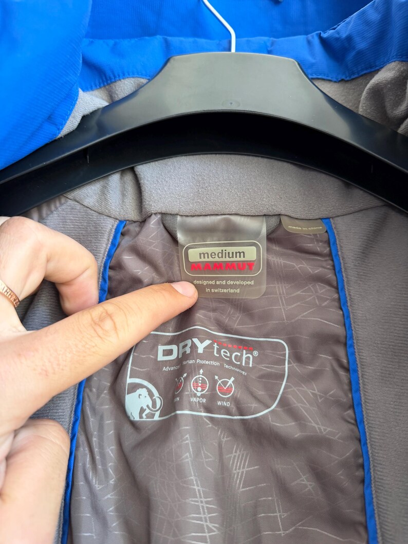 May include: A blue jacket with a grey interior lining featuring the Mammut brand logo and text. The label indicates a size medium, designed and developed in Switzerland. The interior fabric displays Drytech technology branding with icons for rain, vapor, and wind protection.