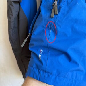 May include: A bright blue jacket with dark gray panels and gray zipper pulls. The jacket features a small white tear in the fabric near the pocket seam, highlighted by a red circle. The text DRYtech is printed in white on the lower front section.