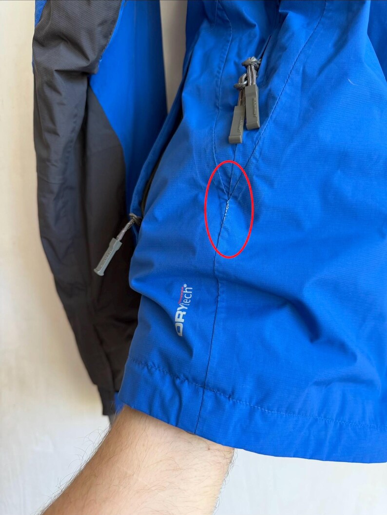 May include: A bright blue jacket with dark gray panels and gray zipper pulls. The jacket features a small white tear in the fabric near the pocket seam, highlighted by a red circle. The text DRYtech is printed in white on the lower front section.