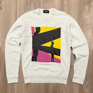 May include: A white sweatshirt featuring a graphic design with black silhouettes against yellow and pink geometric shapes. The sweatshirt has a crew neck and long sleeves. The brand name is visible at the top.