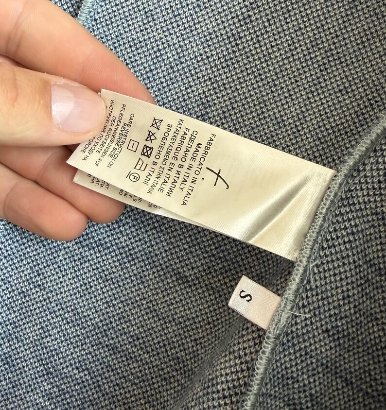 May include: A blue and white textured knit garment featuring a white fabric care label with the text Made in Italy and a small separate white tag with the letter S. The label includes various laundry symbols and multilingual instructions.