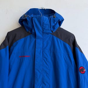 May include: A bright blue hooded jacket with dark gray shoulder panels. The jacket features a front zipper closure, a hood with adjustable drawstrings, and the red Mammut logo text on the chest. A circular patch with a black mammoth icon is on the sleeve.