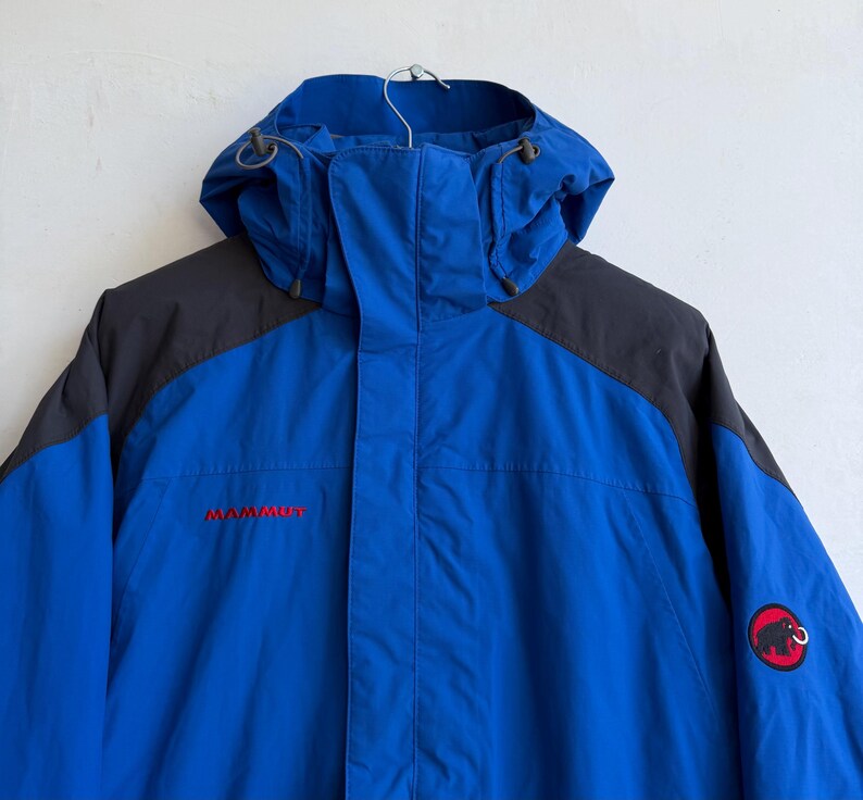 May include: A bright blue hooded jacket with dark gray shoulder panels. The jacket features a front zipper closure, a hood with adjustable drawstrings, and the red Mammut logo text on the chest. A circular patch with a black mammoth icon is on the sleeve.