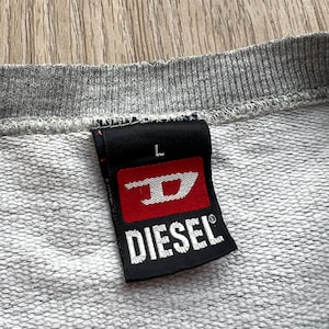 May include: A grey sweatshirt with a black and red Diesel brand label. The label has the Diesel logo and the letter "L" for size.