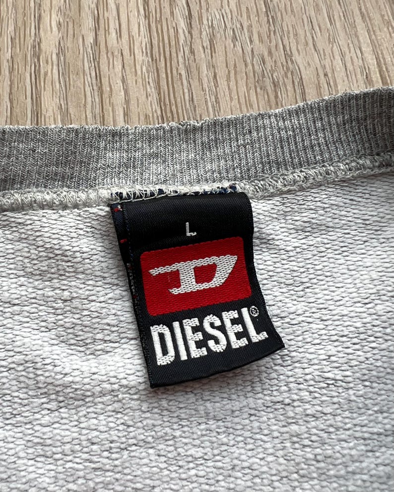 May include: A grey sweatshirt with a black and red Diesel brand label. The label has the Diesel logo and the letter "L" for size.