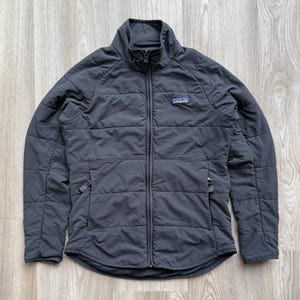 May include: A dark grey quilted jacket with a full-length zip and a stand-up collar. The jacket has a small logo patch on the chest and two side pockets. The jacket is made of a lightweight material, suitable for outdoor activities.