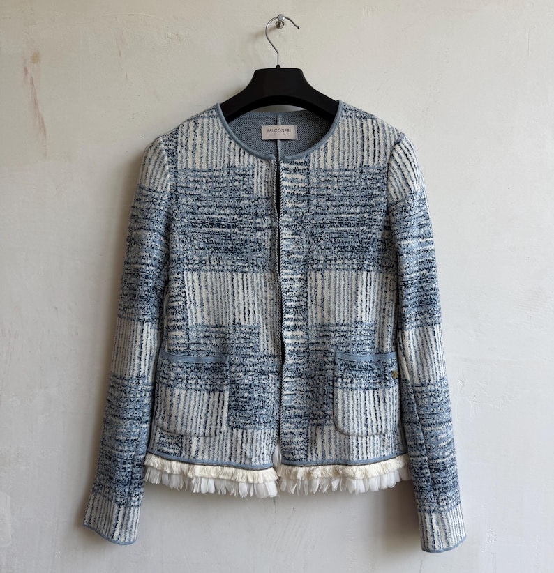 May include: A long-sleeved jacket featuring a blue and white textured grid pattern. The garment has a round neckline, a front zipper closure, two front pockets, and a white fringe trim along the bottom hem. The interior label reads FALCONERI.