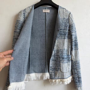 May include: A blue and white open-front jacket featuring a textured, multi-tonal plaid pattern, a round neckline, and long sleeves. The garment includes two front pockets and a white fringed trim along the hem. A label reads FALCONERI.