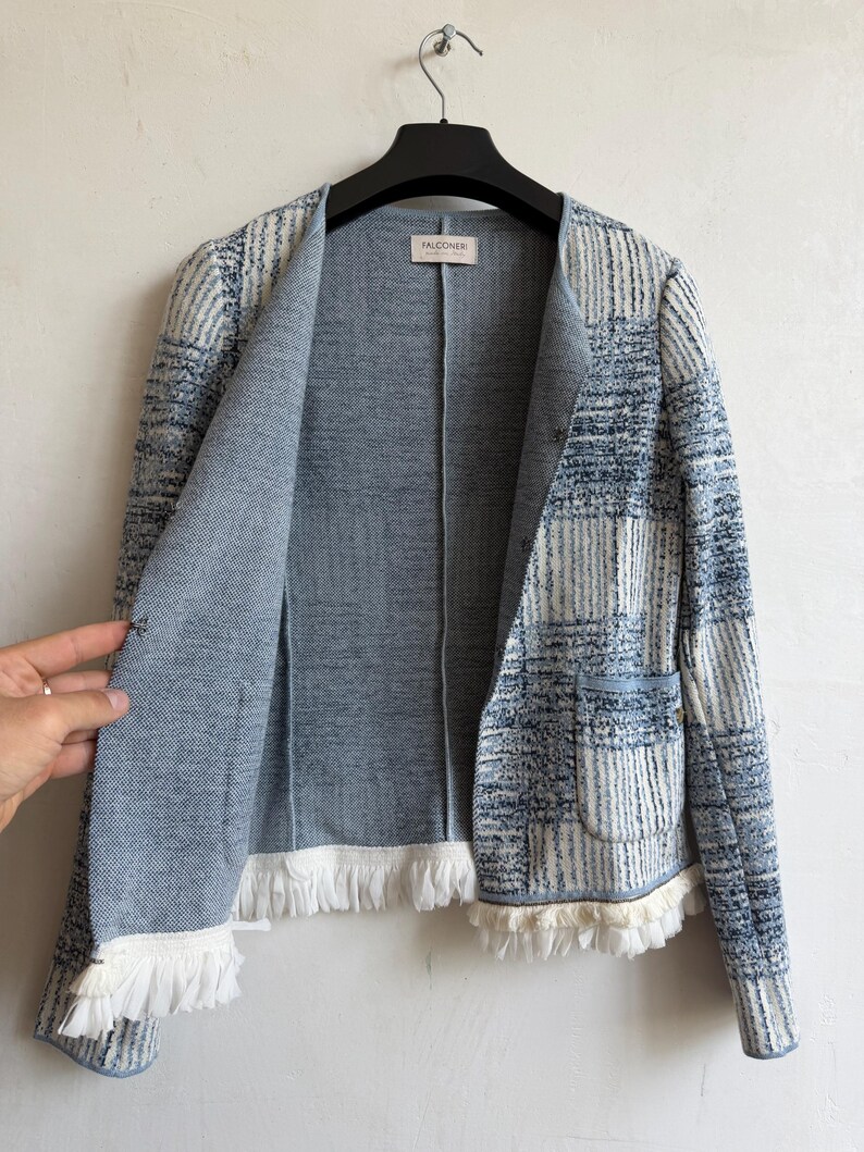 May include: A blue and white open-front jacket featuring a textured, multi-tonal plaid pattern, a round neckline, and long sleeves. The garment includes two front pockets and a white fringed trim along the hem. A label reads FALCONERI.