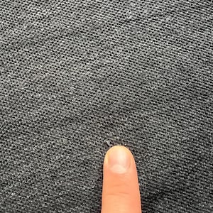 May include: Close-up of a dark gray textured fabric with a small hole in the center. A finger is pointing at the hole.