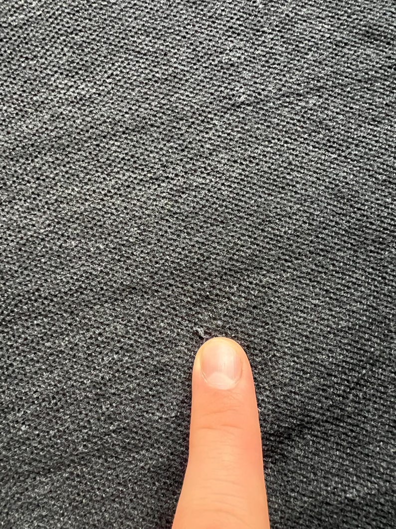 May include: Close-up of a dark gray textured fabric with a small hole in the center. A finger is pointing at the hole.