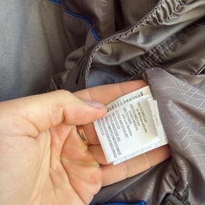 May include: A grey jacket interior featuring a blue trim and a white care label with black text. The label specifies 100% polyamide fabric bonded to a membrane, 100% polyester lining, and 100% polyester padding. A grey drawstring with a toggle is visible.