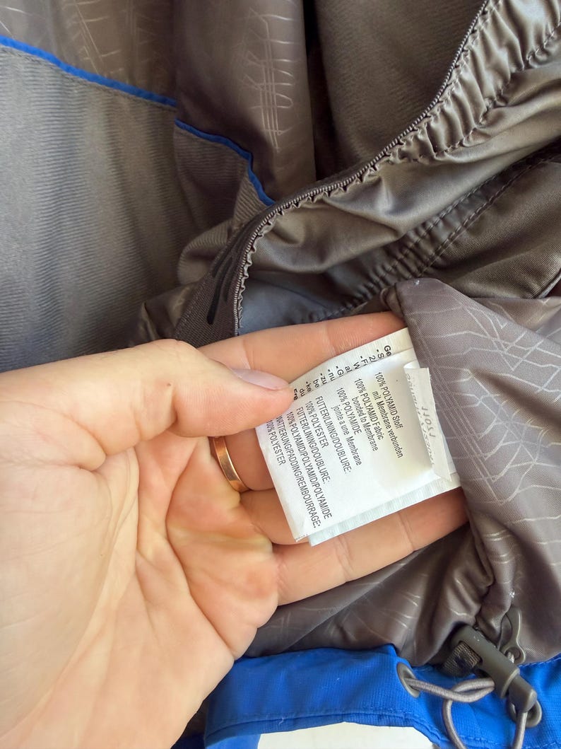 May include: A grey jacket interior featuring a blue trim and a white care label with black text. The label specifies 100% polyamide fabric bonded to a membrane, 100% polyester lining, and 100% polyester padding. A grey drawstring with a toggle is visible.