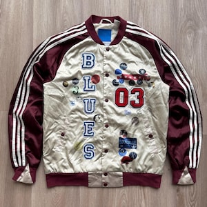 May include: Cream-colored satin bomber jacket with maroon accents and white stripes on the sleeves. The jacket features the word "BLUES" and the number "03" in patches, along with various other decorative patches.
