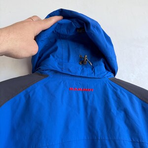 May include: A bright blue hooded jacket with dark gray shoulder panels. The word MAMMUT is embroidered in red on the upper back. The hood features an adjustable drawstring with a plastic toggle, and the fabric has a smooth, technical finish.