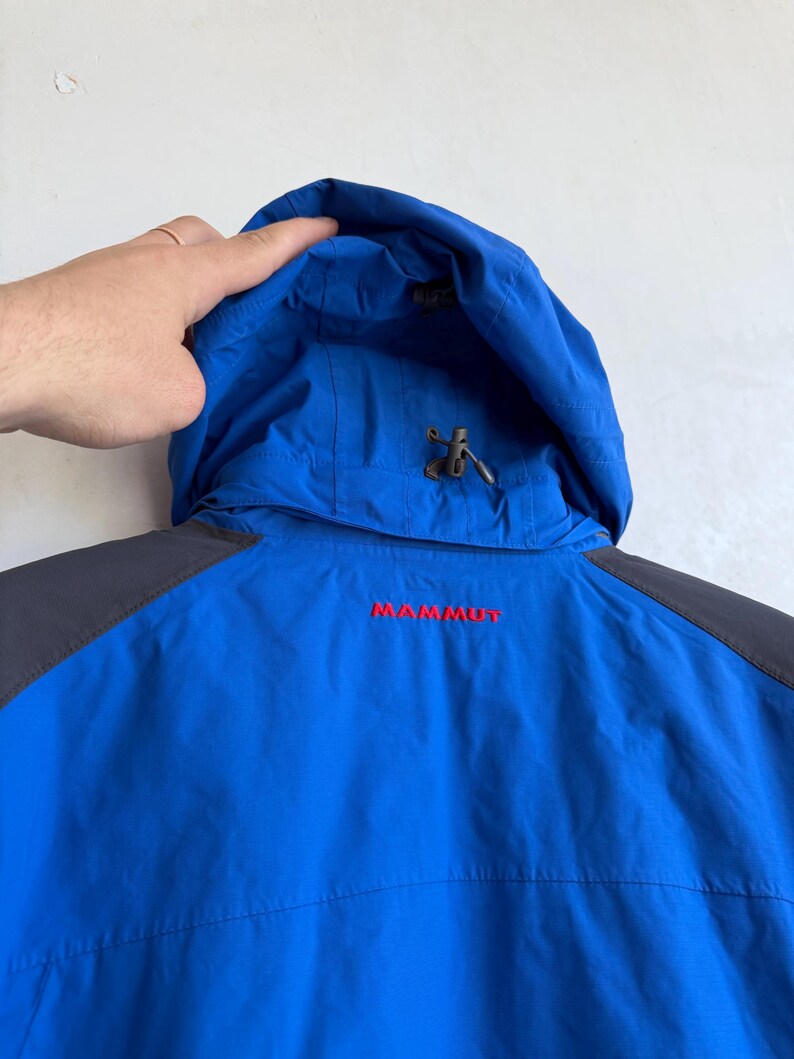 May include: A bright blue hooded jacket with dark gray shoulder panels. The word MAMMUT is embroidered in red on the upper back. The hood features an adjustable drawstring with a plastic toggle, and the fabric has a smooth, technical finish.