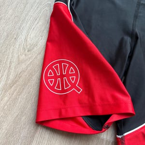 May include: Black and red athletic shorts with a white logo on the red panel. The shorts have a white stripe and stitching detail. The logo is a stylized basketball with the letter "Q".