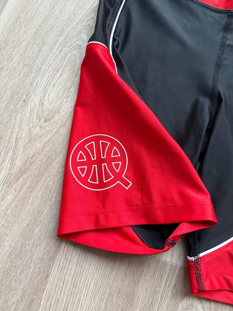 May include: Black and red athletic shorts with a white logo on the red panel. The shorts have a white stripe and stitching detail. The logo is a stylized basketball with the letter "Q".