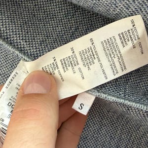 May include: A blue and white textured fabric garment with a white care label attached. The label lists material composition as 100% cotton, 65% polyester, 30% cotton, and 5% copper. A small white tag with the letter S is attached to the seam.