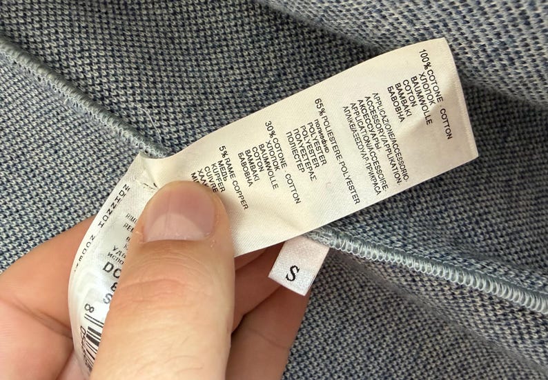 May include: A blue and white textured fabric garment with a white care label attached. The label lists material composition as 100% cotton, 65% polyester, 30% cotton, and 5% copper. A small white tag with the letter S is attached to the seam.