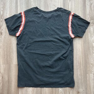 May include: A dark gray t-shirt with white and red stripes on the sleeves.