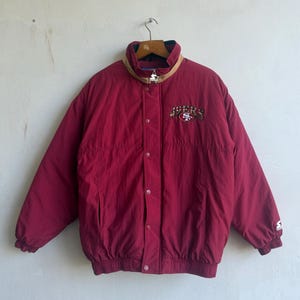May include: A burgundy Starter jacket with a button-up front and a ribbed hem. The jacket features the "49ers" logo embroidered on the chest and a tan collar. The jacket is hanging from a wooden hanger.