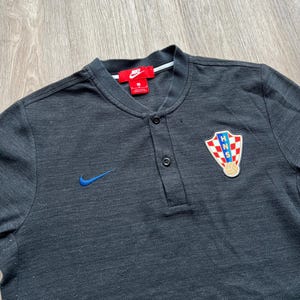May include: Black short-sleeved polo shirt with a blue Nike swoosh logo and a Croatian national team crest. The shirt has a button-up collar and a single button placket.