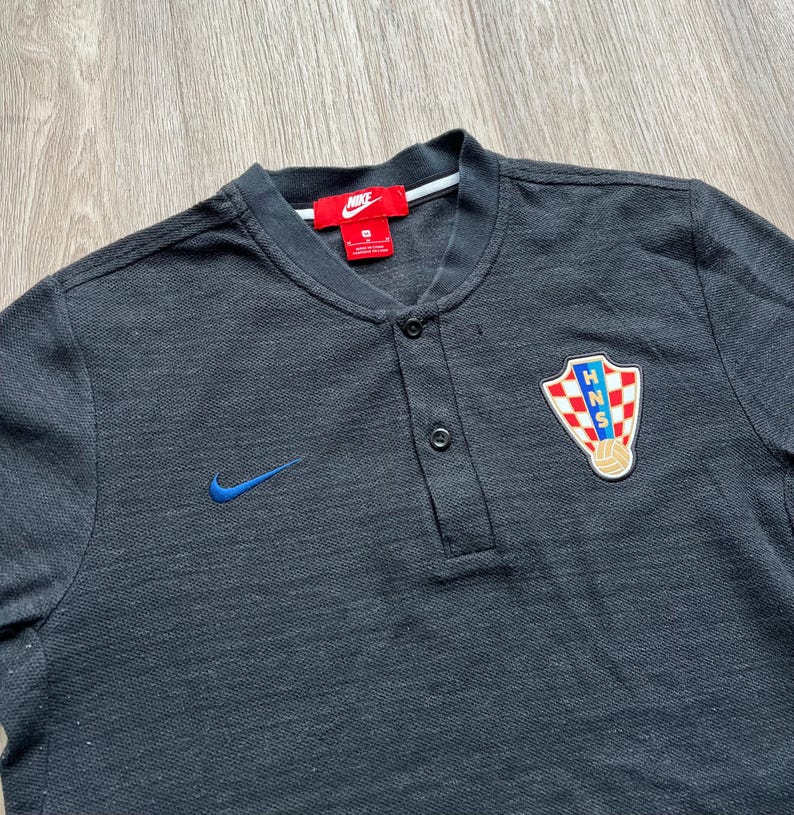 May include: Black short-sleeved polo shirt with a blue Nike swoosh logo and a Croatian national team crest. The shirt has a button-up collar and a single button placket.