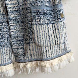 May include: A knit cardigan featuring a blue, white, and dark navy abstract pattern with a textured finish. The garment includes a large patch pocket on the front, a small circular metallic button, and a hemline adorned with white fringe trim.