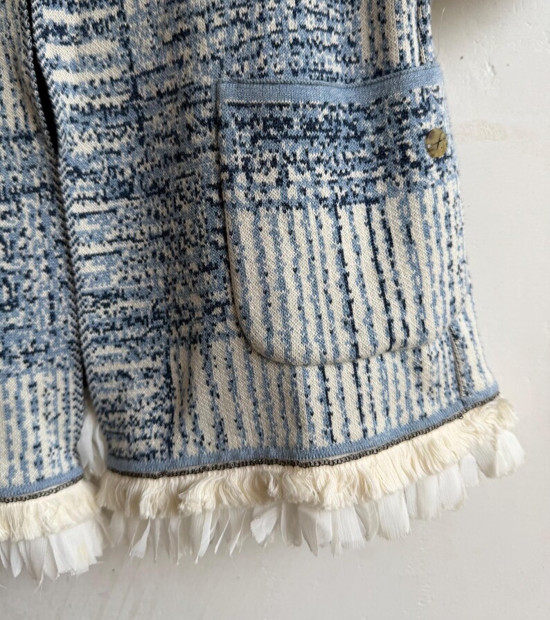 May include: A knit cardigan featuring a blue, white, and dark navy abstract pattern with a textured finish. The garment includes a large patch pocket on the front, a small circular metallic button, and a hemline adorned with white fringe trim.