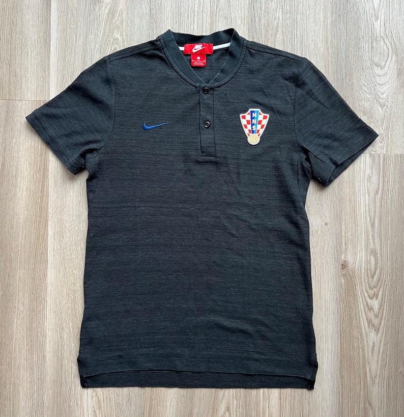 May include: A black short-sleeved polo shirt with a button-up collar. The shirt has a small Nike logo on the left chest and a Croatian national team crest on the right chest.