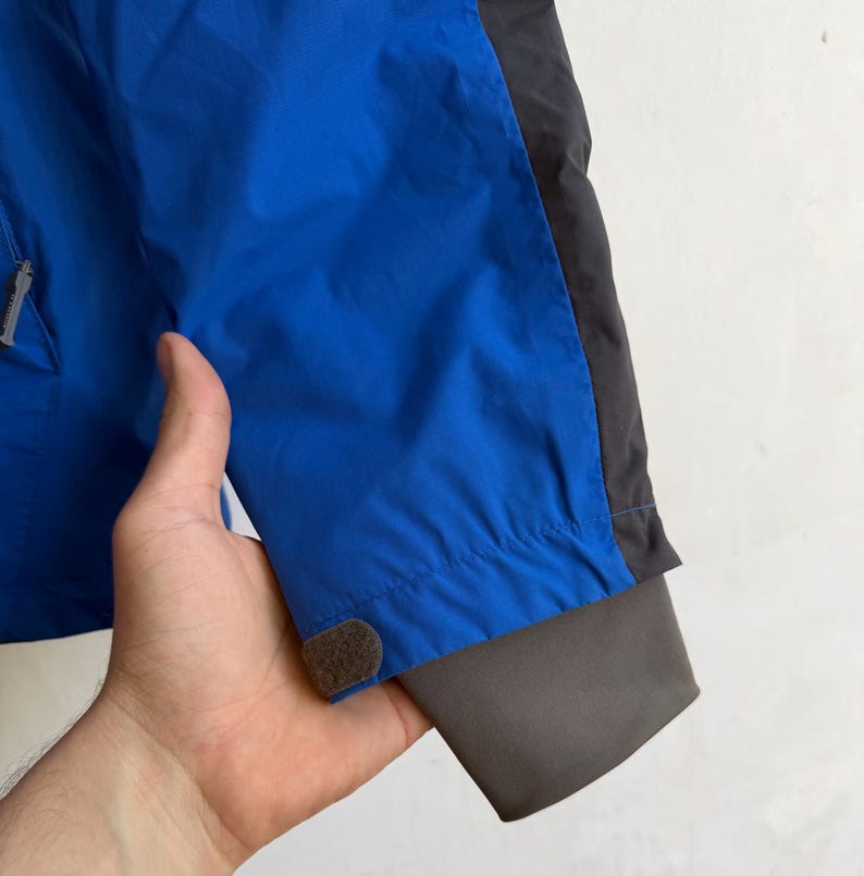 May include: A bright blue jacket sleeve featuring a dark gray side panel, a dark gray inner cuff, and a small hook-and-loop fastener tab at the wrist. The sleeve is constructed from a smooth, synthetic material with a visible zipper pull.