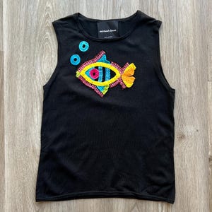 May include: Black sleeveless tank top with a colorful embroidered fish design on the front. The fish has a yellow body, blue and green scales, and a pink eye. The text "michael simon" is printed on the back of the shirt.