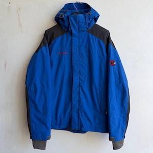 May include: A bright blue hooded jacket with dark gray shoulder and sleeve panels. The jacket features a front zipper closure, a high collar, and adjustable cuffs. A red Mammut logo is embroidered on the left chest and the left upper sleeve.