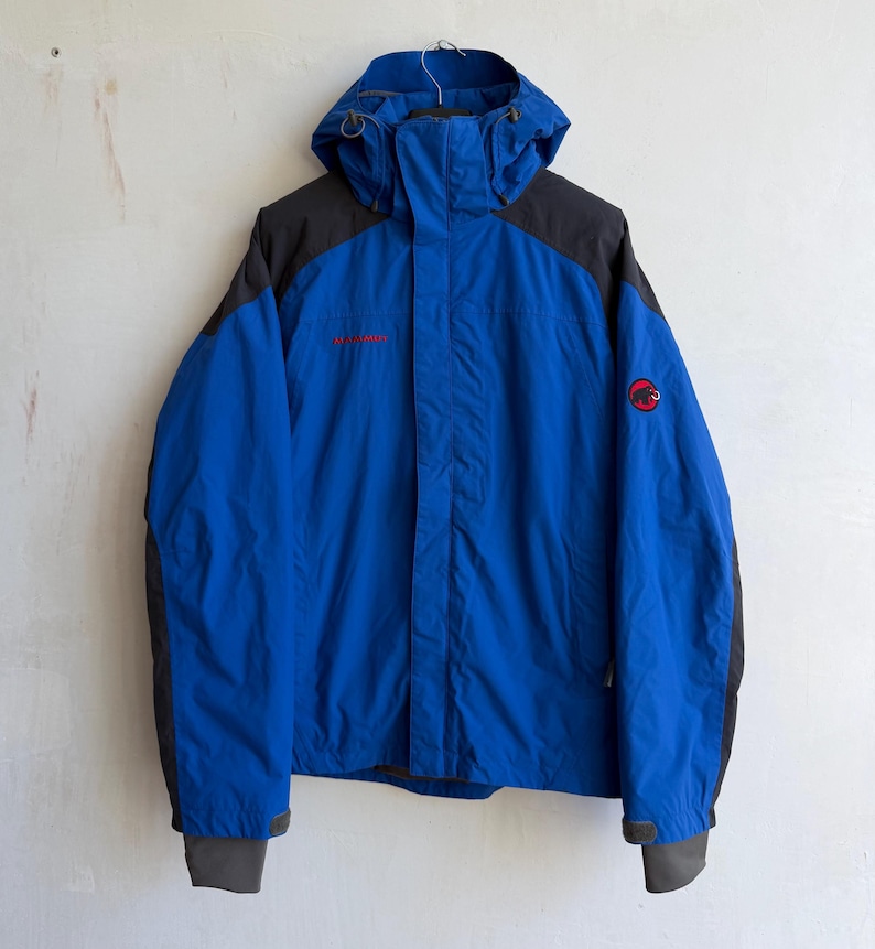 May include: A bright blue hooded jacket with dark gray shoulder and sleeve panels. The jacket features a front zipper closure, a high collar, and adjustable cuffs. A red Mammut logo is embroidered on the left chest and the left upper sleeve.