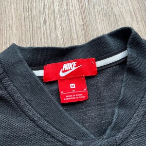May include: A black Nike polo shirt with a red Nike logo and a white tag that says "Made in China, Fabrique en Chine". The shirt has a button closure and a size tag that says "M".