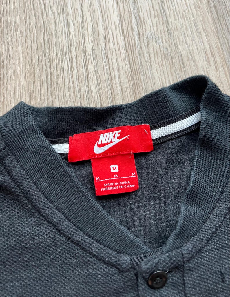 May include: A black Nike polo shirt with a red Nike logo and a white tag that says "Made in China, Fabrique en Chine". The shirt has a button closure and a size tag that says "M".