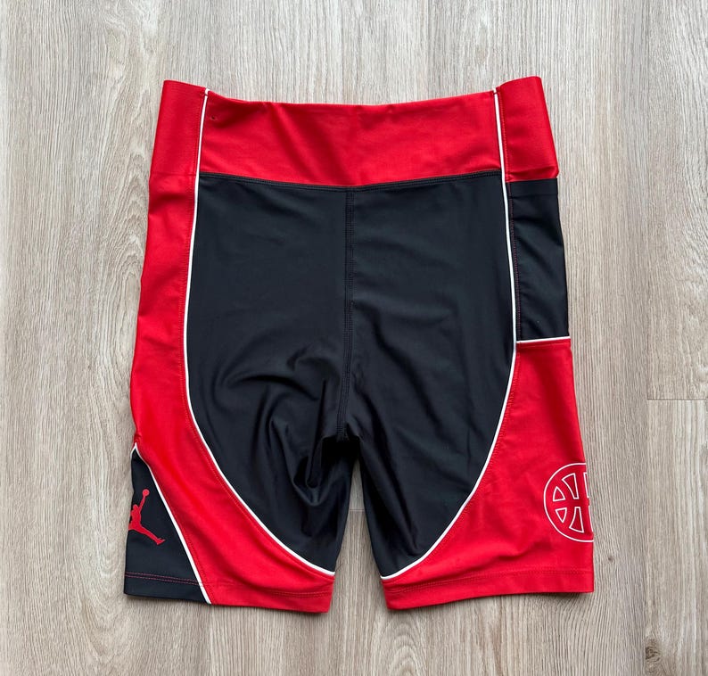 May include: Black and red athletic shorts with white trim. The shorts have a red waistband and side panels, with a black center. The Jordan logo is visible on the left leg, and a circular design on the right leg.