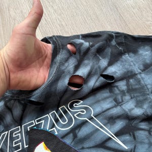 Może przedstawiać: Czarna i szara koszulka tie-dye z dziurami w pobliżu kołnierza. Koszulka zawiera napis "YEEZUS" białymi literami i grafikę ponurego żniwiarza.