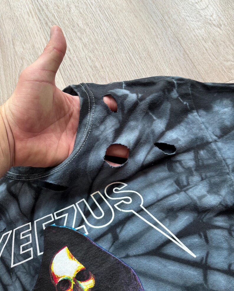 Może przedstawiać: Czarna i szara koszulka tie-dye z dziurami w pobliżu kołnierza. Koszulka zawiera napis "YEEZUS" białymi literami i grafikę ponurego żniwiarza.