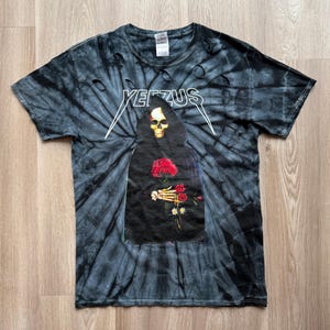 Może przedstawiać: Czarna i szara koszulka tie-dye z napisem "YEEZUS" w kolorze białym. Koszulka przedstawia grafikę szkieletu w czarnej szacie trzymającego czerwone r&oacute;że. Koszulka ma postrzępione detale.