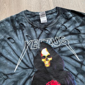 Może przedstawiać: Czarna i szara koszulka tie-dye z postrzępionymi detalami. Koszulka zawiera napis "YEEZUS" w kolorze białym, a także grafikę ponurego żniwiarza z ż&oacute;łtą czaszką i czerwonym sercem. Koszulka ma okrągły dekolt.