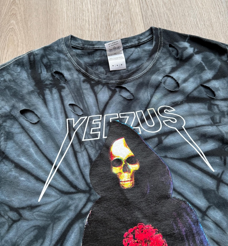 Może przedstawiać: Czarna i szara koszulka tie-dye z postrzępionymi detalami. Koszulka zawiera napis "YEEZUS" w kolorze białym, a także grafikę ponurego żniwiarza z ż&oacute;łtą czaszką i czerwonym sercem. Koszulka ma okrągły dekolt.