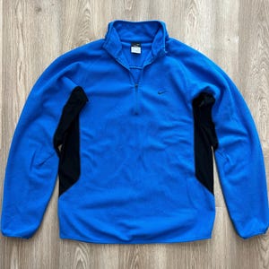 Puede incluir: Un jersey de forro polar azul con cuello alzado con cremallera y paneles laterales negros. El logotipo de Nike es visible en el pecho. La parte superior de manga larga está hecha de un material suave.