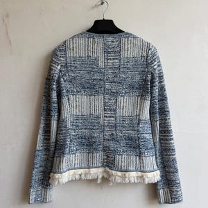 May include: A long-sleeved jacket featuring a blue and white textured plaid pattern. The garment has a round neckline, a straight hemline, and is finished with a decorative fringe trim along the bottom edge. The jacket is displayed on a hanger.